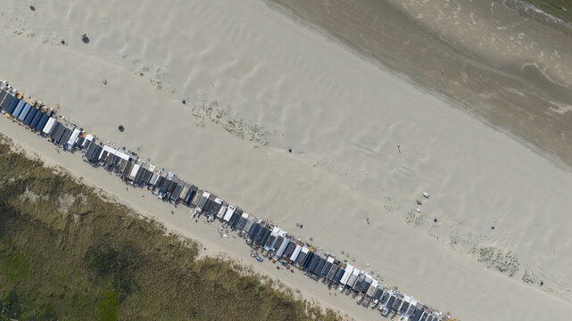 Aerial view of a long row of beach huts and cabins stretching across a wide sandy beach next to grassy dunes in Velsen-Noord, North Holland, Netherlands.