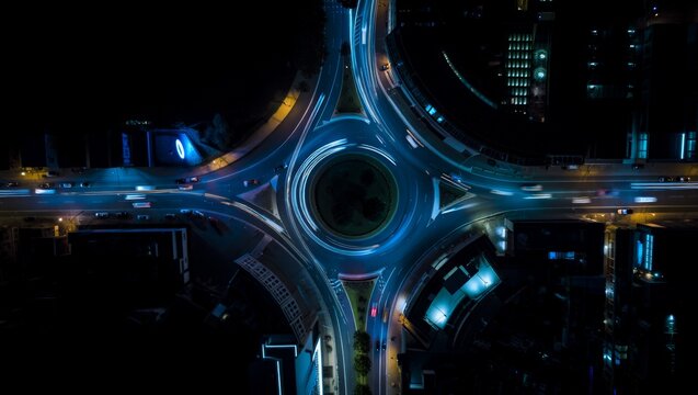 Stunning aerial drone shot looking directly down at a large illuminated roundabout at night, featuring sweeping blue and gold vehicle light trails circling in perfect geometric patterns.