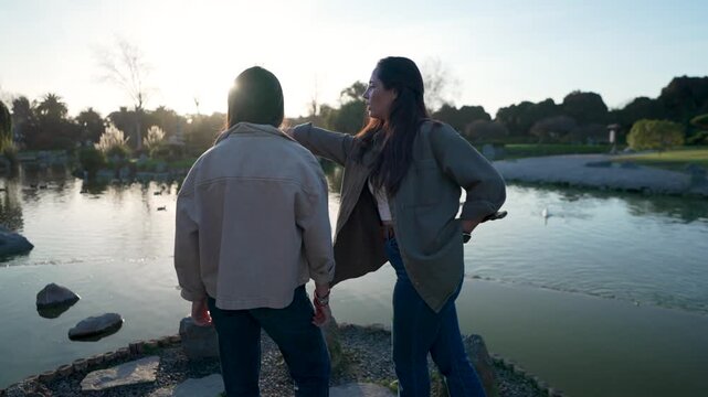 Medium shot of a latina mother talking and pointing at something while standing with her teenage daughter by a park pond