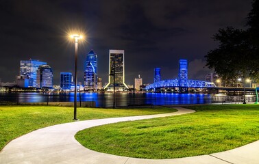 Obraz premium Vibrant night view of downtown Jacksonville, Florida, showcasing the illuminated city skyline, Main Street Bridge, and reflections on the Saint Johns River with a scenic park path