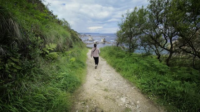 Lone woman walking down a dirt path surrounded by lush green vegetation leading to the ocean with unique rock formations, representing a journey of discovery and solitude in a natural setting