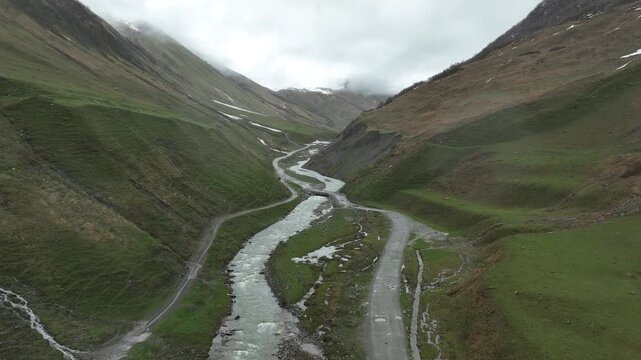 High-angle aerial view capturing a clean, rushing mountain creek flowing from a melting snow patch down a vibrant green alpine valley with rocky terrain and small wildflowers in Ushguli, Georgia