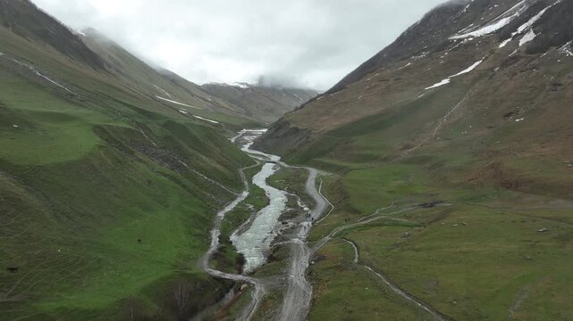 High-angle aerial view capturing a clean, rushing mountain creek flowing from a melting snow patch down a vibrant green alpine valley with rocky terrain and small wildflowers in Ushguli, Georgia