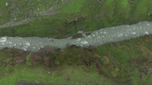 High-angle aerial view capturing a clean, rushing mountain creek flowing from a melting snow patch down a vibrant green alpine valley with rocky terrain and small wildflowers in Ushguli, Georgia
