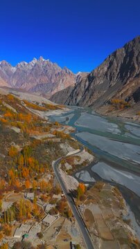 Aerial view of the winding road through Gulmit village with autumn trees and the Hunza River under a clear sky in Hunza Nagar, Gilgit Baltistan, Pakistan.