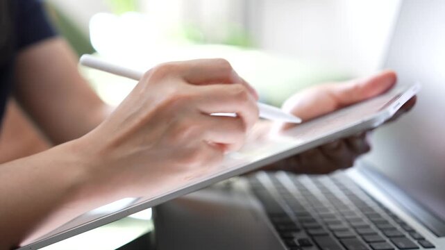 Hand using stylus on tablet beside laptop for electronic signature, digital document review, online approval, paperless workflow, business productivity, remote work, modern technology.