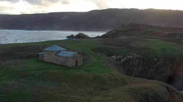 Aerial view of Ermita de Santa Comba chapel on a grassy cliff overlooking the beach and rugged coastline during sunset in Covas, La Coruna, Spain.