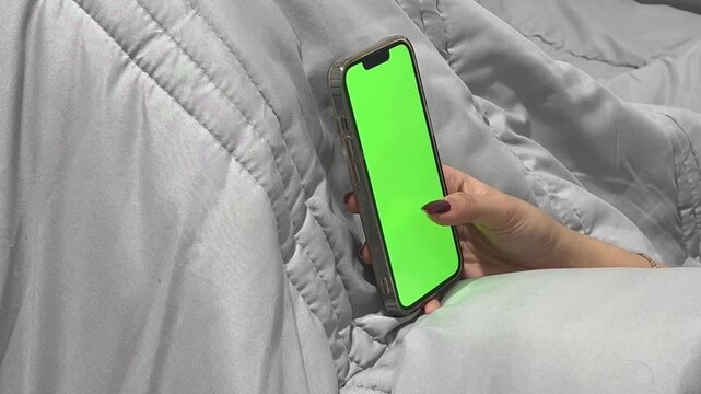 Person scrolling on smartphone with green screen in bed, 16:9 horizontal video, night time habit, bedtime routine, browsing social media, mobile technology, cozy environment.
