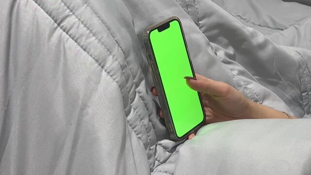 Person scrolling on smartphone with green screen in bed, 16:9 horizontal video, night time habit, bedtime routine, browsing social media, mobile technology, cozy environment.