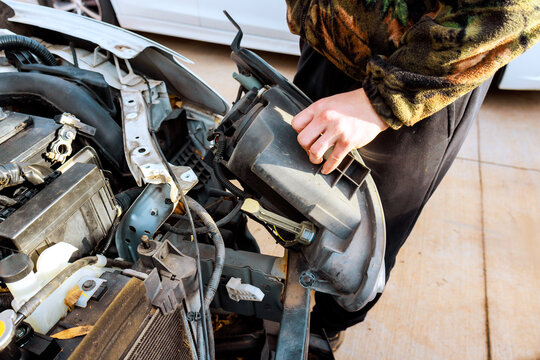 Worker is seen taking part car headlight in garage on near air filter. other parts are visible around him.