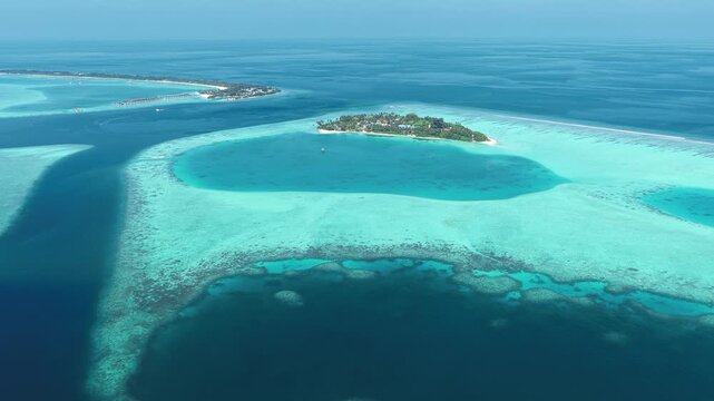 Aerial view of a lush tropical island surrounded by vibrant coral reefs and turquoise lagoons under a clear blue sky in Kudahuvadhoo, Dhaalu Atoll, Maldives.