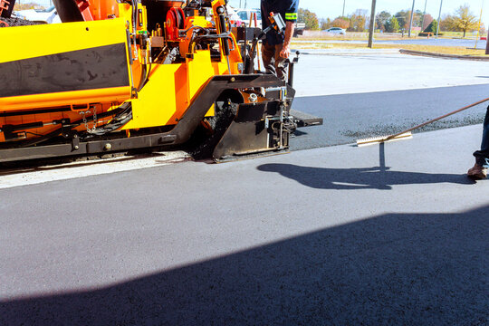 Workers use asphalt paving machines to lay asphalt on parking lot while another worker smooths surface with tool.