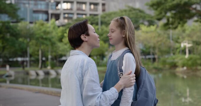 Portrait of happy mother kisses little daughter and sets backpack in autumn park. Woman cares of girl junior student ready for school in public garden