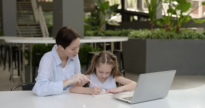 Portrait of attentive female teacher gives explanations to little girl at laptop in office. Mother checks daughter writing in notebook and looks at computer