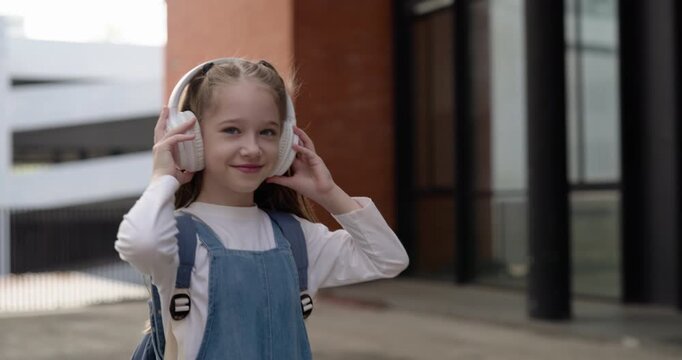 Portrait of junior schoolgirl listens to music via headphones and smiles on street. Cheerful little girl with backpack dances and walks on way to school