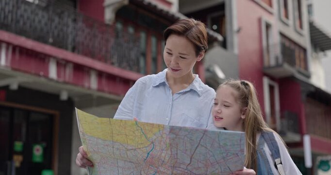 Portrait of woman with little daughter reads map against old buildings. Happy family of mother and girl visits foreign country on holiday. Travel with children