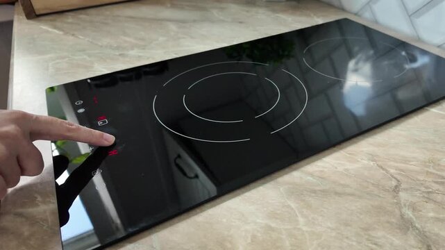 A close-up of a modern black induction cooktop with a hand adjusting the settings. The surface is sleek and shiny, reflecting light.