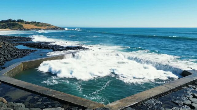 Ocean waves crashing over a hexagonal saltwater swimming pool near the shoreline on a sunny day