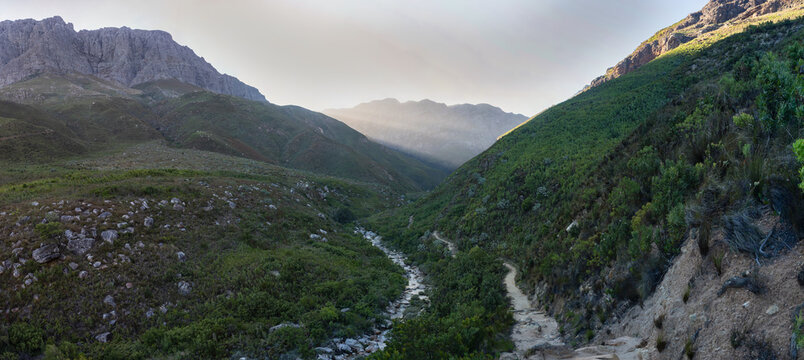 High resolution stitched panorama of Jonkershoek Nature Reserve, South Africa, featuring a winding hiking trail by the Eerste River valley beautifully unevenly lit by dramatic morning sunrise rays.