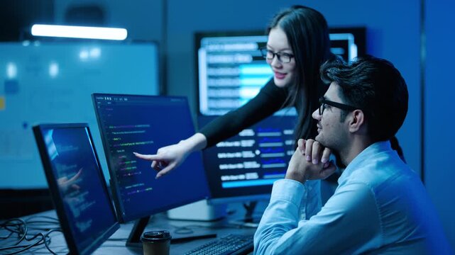 Two software engineers collaborate on computer coding projects in a high-tech office at night. A female programmer points to a screen, discussing application development with her male colleague.