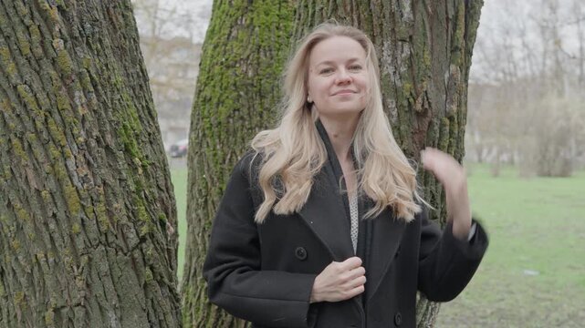 Young beautiful woman in black coat standing by trees and fixing hair