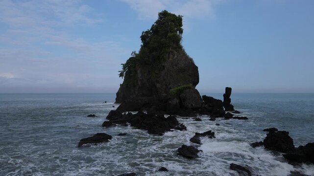 Aerial view of Pantai Karang Agung Kebumen, a majestic sea stack topped with lush greenery surrounded by dark volcanic rocks and crashing ocean waves in Ayah, Central Java, Indonesia.