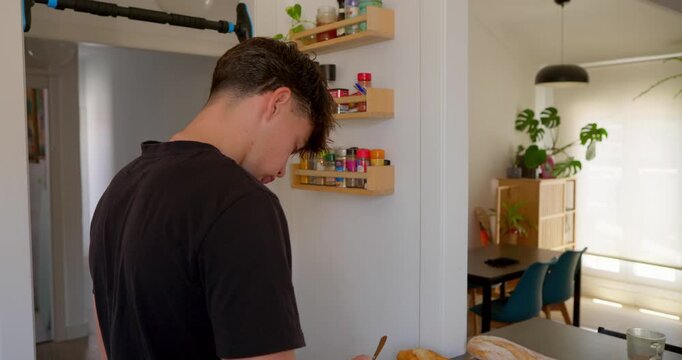 Young man in kitchen: A young man with trendy haircut and black t-shirt leans over the kitchen counter, deep in thought while preparing food.
