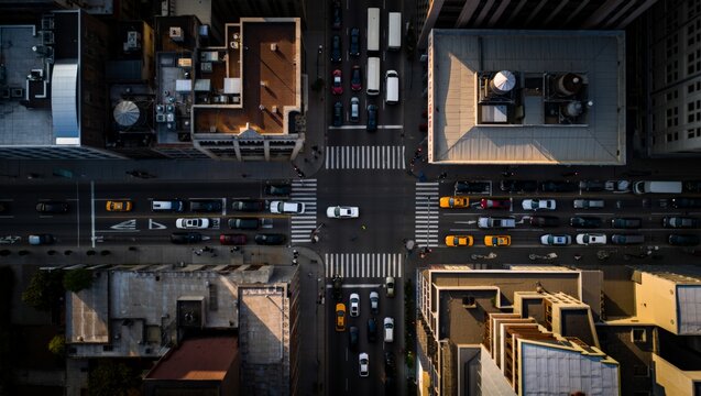Directly overhead aerial view of a busy urban intersection at warm sunset showing yellow taxis, trucks, pedestrians on crosswalks, long shadows, and building rooftop details.