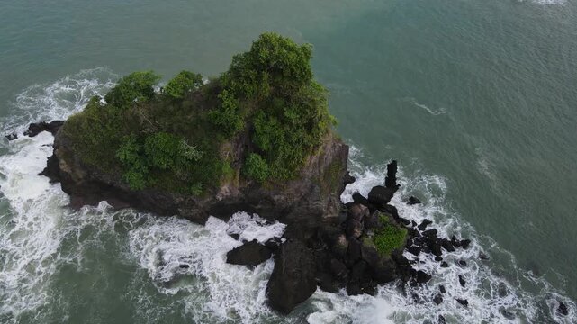 Aerial view of Pantai Karang Agung Kebumen featuring a rocky island covered in lush green trees surrounded by crashing white waves in Ayah, Central Java, Indonesia.