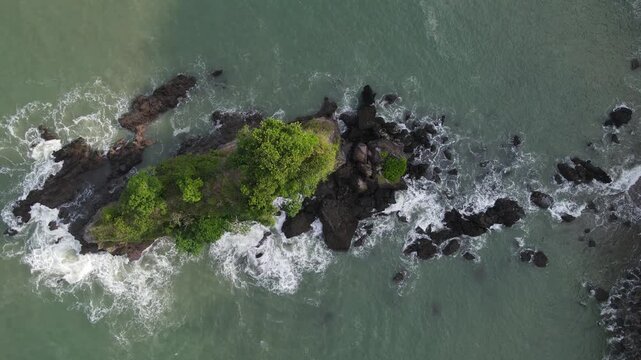 Aerial view of the rocky islet at Karang Agung Beach with green trees and waves crashing against the shore in Ayah, Central Java, Indonesia.