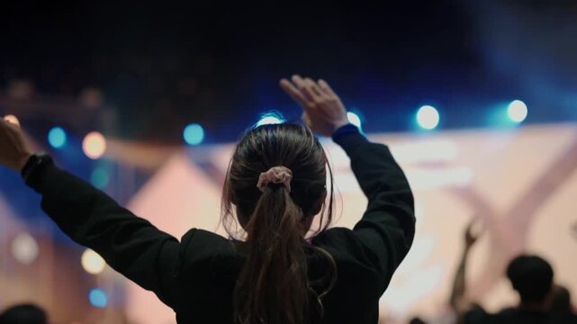 Rear view of a woman raising her hands during a live worship concert. Blurred stage lights and crowd in the background create an emotional atmosphere of faith, spirituality, hope, inspiration, and com