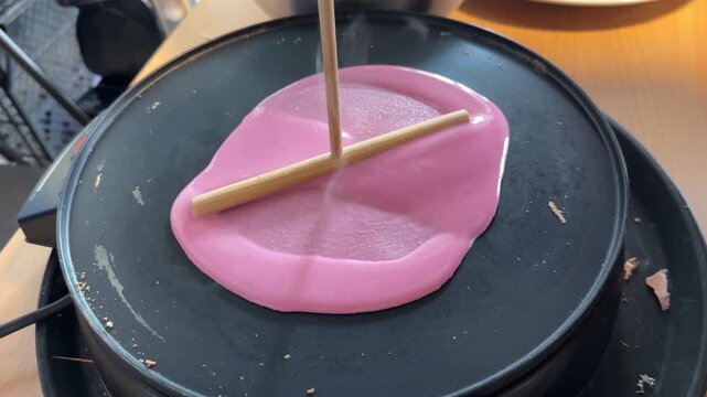 Spreading pink pancake batter with a wooden T-shaped crepe spreader tool on a hot electric griddle plate