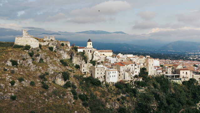 A scenic panoramic view of the historic old town of Scalea in Calabria, Italy. Ancient castle ruins stand on a rocky hill overlooking stone houses and distant mountains under clouds.