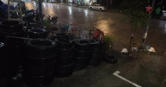 At night, rain slicked tire stacks sit outside a mechanic's shop on a street, while vehicles traverse the shimmering, drenched asphalt road under a heavy downpour.