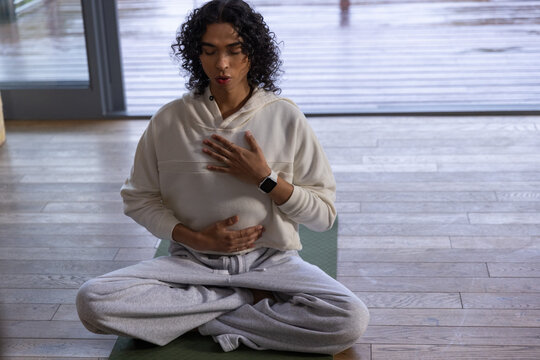 Non-binary person meditating cross-legged on green mat by glass door, wearing cream hoodie
