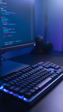 Low angle view of illuminated mechanical keyboard and code editor monitor screen in moody dark office, programming and software development vertical photo