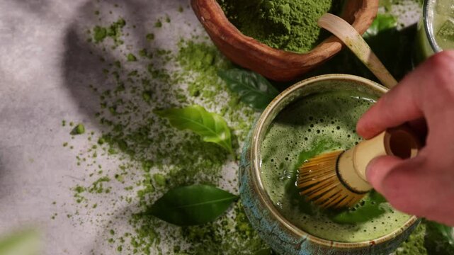 Hand whisking vibrant green matcha tea with traditional bamboo whisk, surrounded by matcha powder and tea-making ingredients. Illuminated by natural sunlight, creating an authentic tea ceremony scene