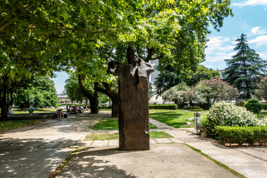 Fan Noli statue in Tirana, Albania, stands prominently in a green park shaded by large trees, with a stone walkway and a fountain in the background.