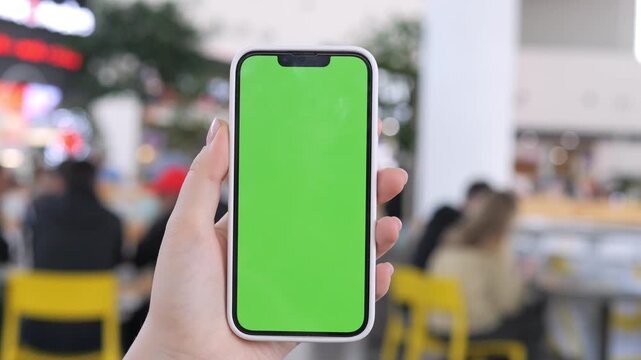Hand holds phone with green screen in lively indoor cafe. Blurred background shows people seated at tables. Useful for app demos, marketing, or UI design projects