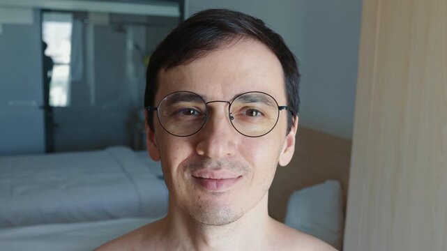 Portrait of a cheerful young adult man wearing stylish eyeglasses, looking at the camera and smiling sincerely in a modern apartment. His positive emotion conveys confidence and contentment