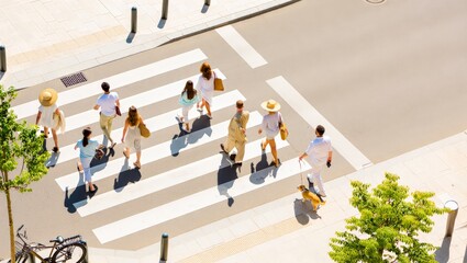 Obraz premium A bustling city street scene captured from above, showing people of various ages and styles crossing the road at a zebra crossing, surrounded by trees, a bicycle, and urban infrastructure.