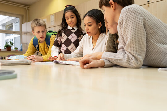 A small group of students is in their elementary classroom while a mixed-race teacher guides them through a math and geometry lesson. She is asking questions and helping them stay focused as they work