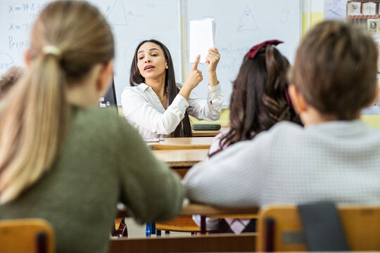 A small group of students is in their elementary classroom while a mixed-race teacher guides them through a math and geometry lesson. She is asking questions and helping them stay focused.