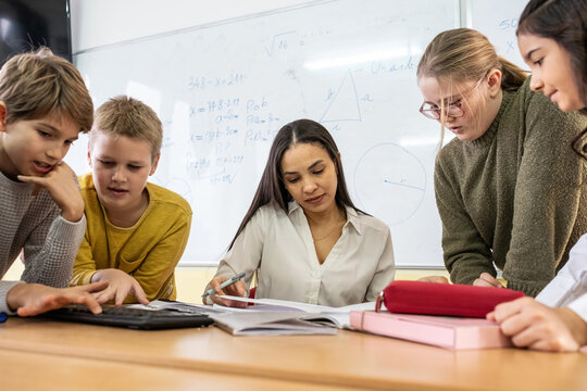 A small group of students is in their elementary classroom while a mixed-race teacher guides them through a math and geometry lesson. She is asking questions and helping them stay focused as they work