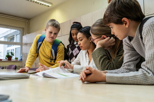 A small group of students is in their elementary classroom while a mixed-race teacher guides them through a math and geometry lesson. She is asking questions and helping them stay focused as they work