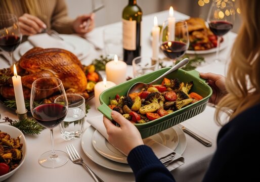 Closeup of a person serving roasted vegetables at a festive holiday dinner table with a roasted turkey, wine, and candles