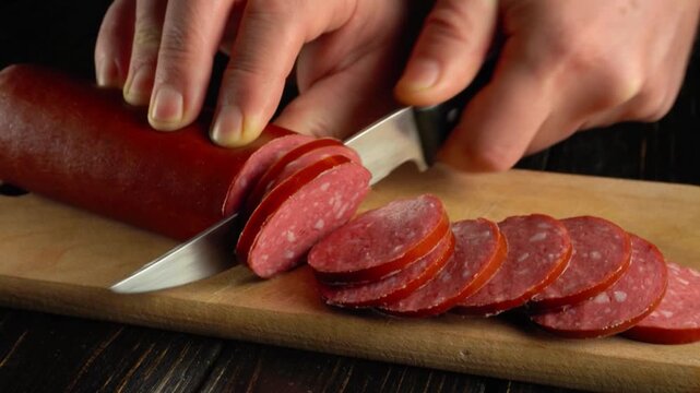 Hands slicing sausage into thin rounds on a wooden cutting board with a sharp knife, showcasing a continuous action of food preparation
