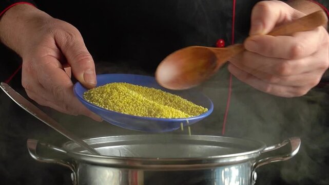 Chef pours yellow couscous from a blue bowl into a stainless steel pot, steam rising, showcasing the cooking process in a kitchen setting