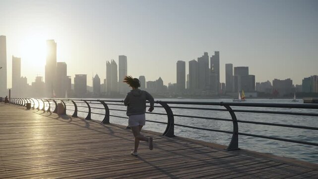 A young East Asian woman wearing casual activewear jogs along a seaside city promenade during early morning golden hour, with urban skyline across the water.
