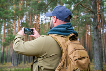 Fototapeta premium traveler with backpack uses smartphone to photograph pine woodland during hike. Nature photography and outdoor adventure concept